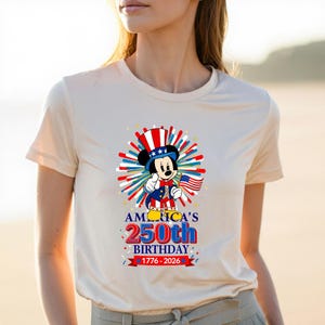 May include: A cream-colored t-shirt featuring a patriotic Mickey Mouse design. Mickey is dressed in a red, white, and blue outfit, holding an American flag. The shirt also displays the text "AMERICA'S 250th BIRTHDAY 1776-2026".