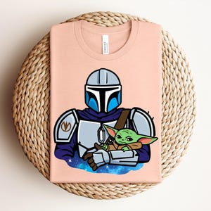 Puede incluir: Una camiseta de color melocot&oacute;n claro con una ilustraci&oacute;n de dibujos animados de un personaje con armadura plateada y azul que sostiene una peque&ntilde;a criatura verde. El dise&ntilde;o est&aacute; sobre un fondo estrellado azul y morado. La camiseta est&aacute; doblada sobre una superficie tejida.