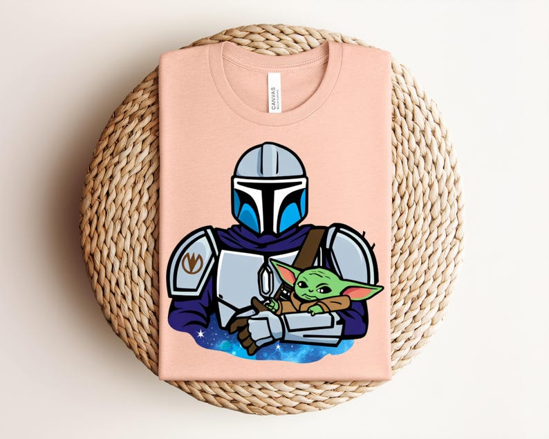 Puede incluir: Una camiseta de color melocot&oacute;n claro con una ilustraci&oacute;n de dibujos animados de un personaje con armadura plateada y azul que sostiene una peque&ntilde;a criatura verde. El dise&ntilde;o est&aacute; sobre un fondo estrellado azul y morado. La camiseta est&aacute; doblada sobre una superficie tejida.