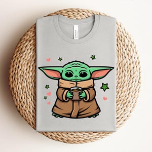 May include: A light gray t-shirt featuring a cartoon illustration of a green alien character holding a cup. The design includes green stars and pink hearts. The shirt is displayed on a woven, circular placemat.