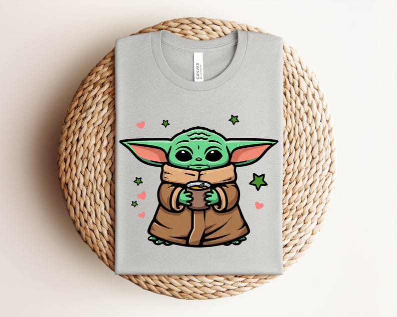 May include: A light gray t-shirt featuring a cartoon illustration of a green alien character holding a cup. The design includes green stars and pink hearts. The shirt is displayed on a woven, circular placemat.