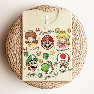 May include: A light tan t-shirt featuring Super Mario characters: Daisy, Mario, Princess Peach, Luigi, Yoshi, and Toad. The shirt includes the text "Super Mario" and character names. The design incorporates stars, mushrooms, and coins.