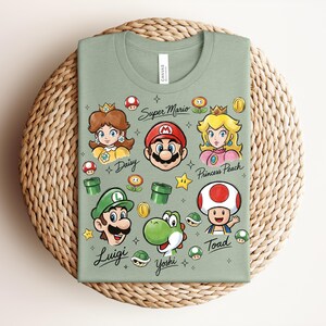 May include: Sage green t-shirt featuring colorful illustrations of Super Mario characters, including Mario, Luigi, Princess Peach, and Yoshi. The shirt has the text "Super Mario" and character names. The shirt is folded on a woven mat.