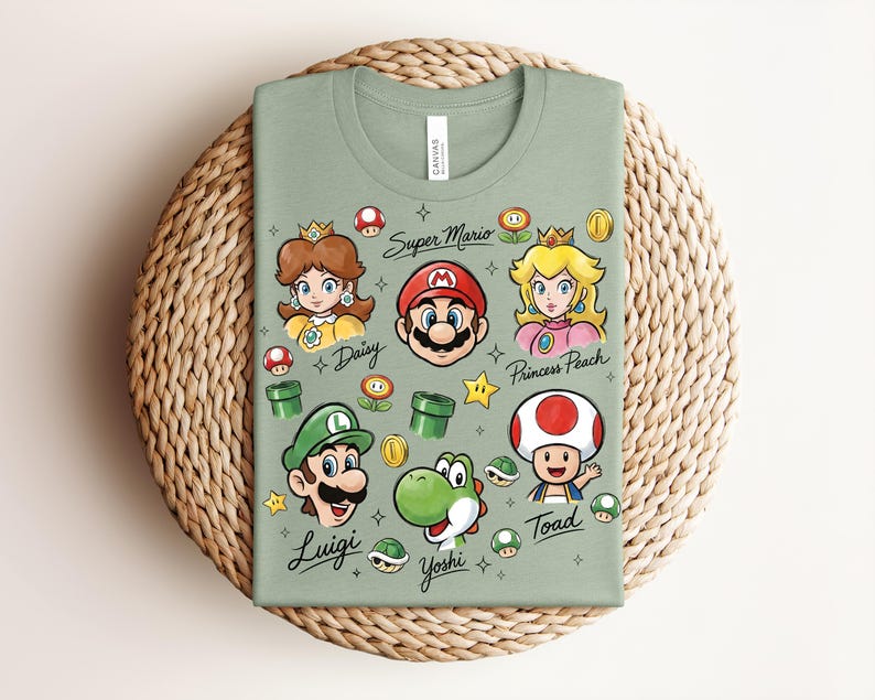 May include: Sage green t-shirt featuring colorful illustrations of Super Mario characters, including Mario, Luigi, Princess Peach, and Yoshi. The shirt has the text "Super Mario" and character names. The shirt is folded on a woven mat.