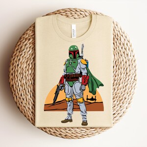 May include: Beige t-shirt featuring a graphic of Boba Fett from Star Wars. The illustration shows Boba Fett in his green, gray, and red armor, with a blaster and cape. The background includes a sunset and a silhouette of a spaceship.