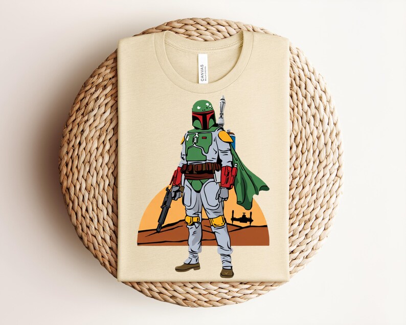 May include: Beige t-shirt featuring a graphic of Boba Fett from Star Wars. The illustration shows Boba Fett in his green, gray, and red armor, with a blaster and cape. The background includes a sunset and a silhouette of a spaceship.