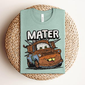 May include: A light green t-shirt featuring a graphic illustration of the character Mater from the movie Cars. The design shows the rusty brown tow truck with large eyes, a wide smile, and the text MATER printed in bold white letters above it.