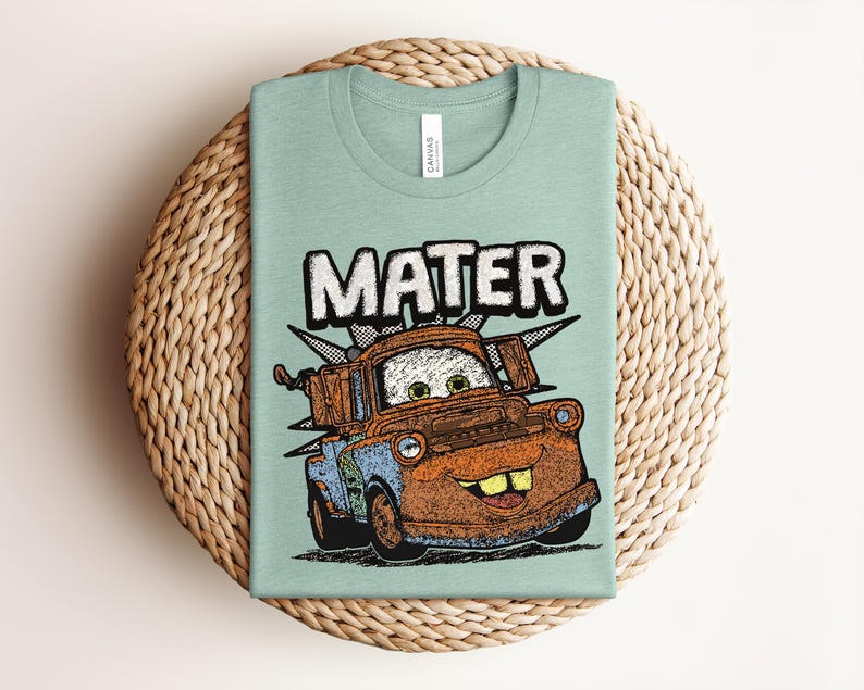 May include: A light green t-shirt featuring a graphic illustration of the character Mater from the movie Cars. The design shows the rusty brown tow truck with large eyes, a wide smile, and the text MATER printed in bold white letters above it.