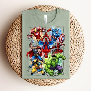 May include: A sage green t-shirt with a vibrant graphic of Marvel superheroes. The design includes Iron Man, Captain America, Thor, Spider-Man, Wolverine, Deadpool, and the Hulk, with watercolour accents. The image is set within a grid.