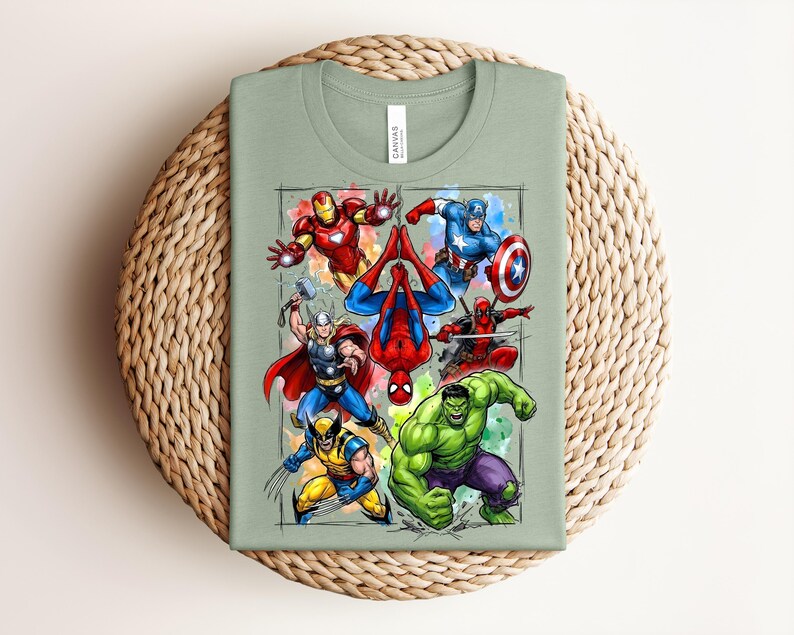 May include: A sage green t-shirt with a vibrant graphic of Marvel superheroes. The design includes Iron Man, Captain America, Thor, Spider-Man, Wolverine, Deadpool, and the Hulk, with watercolour accents. The image is set within a grid.