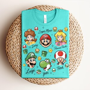 May include: A turquoise t-shirt featuring cartoon illustrations of Super Mario characters, including Mario, Luigi, Princess Peach, and Yoshi. The shirt has the text "Super Mario" and character names. The shirt is folded on a woven surface.