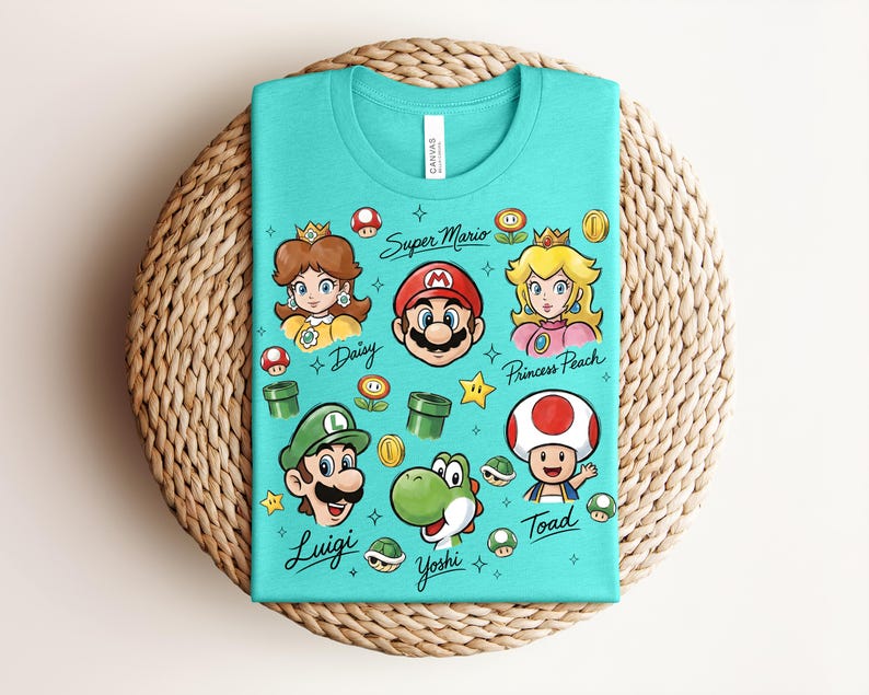 May include: A turquoise t-shirt featuring cartoon illustrations of Super Mario characters, including Mario, Luigi, Princess Peach, and Yoshi. The shirt has the text "Super Mario" and character names. The shirt is folded on a woven surface.