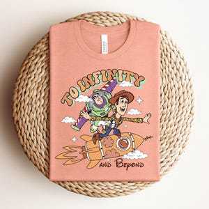 May include: A peach-colored t-shirt featuring a graphic print of Buzz Lightyear and Woody from Toy Story riding a rocket. The text "TO INFINITY" is arched above the characters, and "AND BEYOND" is printed below the rocket, surrounded by clouds.