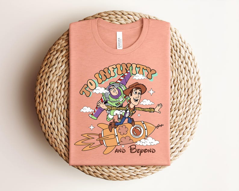 May include: A peach-colored t-shirt featuring a graphic print of Buzz Lightyear and Woody from Toy Story riding a rocket. The text "TO INFINITY" is arched above the characters, and "AND BEYOND" is printed below the rocket, surrounded by clouds.