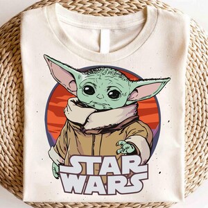 May include: Cream-colored t-shirt featuring a graphic of Grogu, also known as Baby Yoda, from Star Wars. The design includes a green-skinned character with large ears, a tan coat, and the Star Wars logo in white. The background is a red and orange circle.