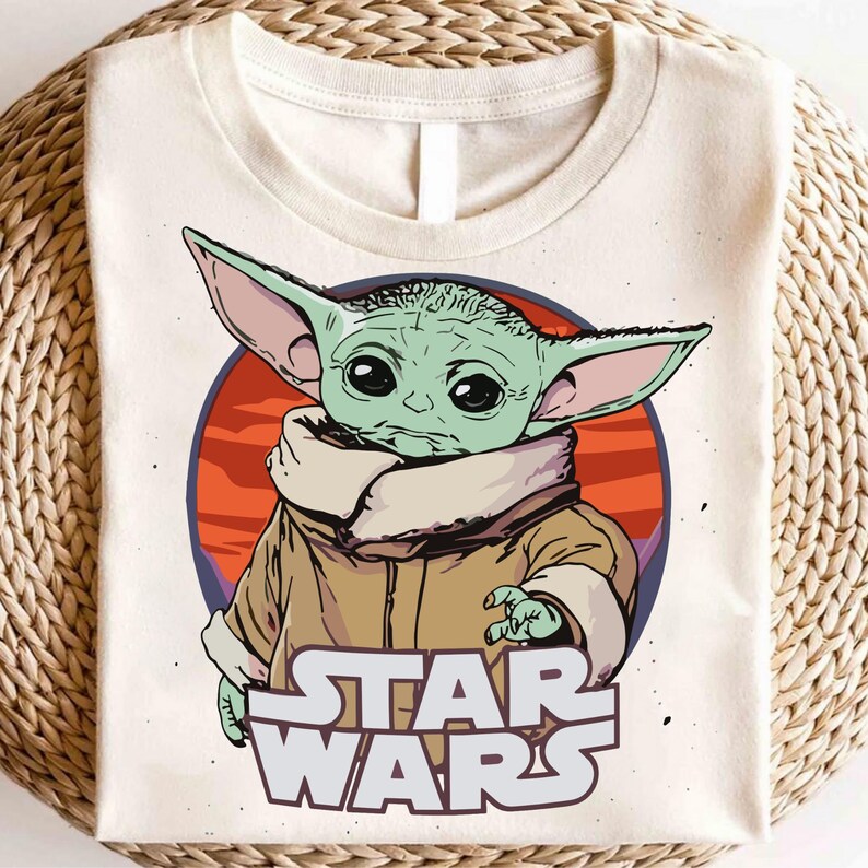 May include: Cream-colored t-shirt featuring a graphic of Grogu, also known as Baby Yoda, from Star Wars. The design includes a green-skinned character with large ears, a tan coat, and the Star Wars logo in white. The background is a red and orange circle.