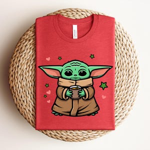 May include: Red t-shirt featuring a cartoon illustration of a green alien character holding a cup. The character is wearing a brown coat and scarf, with green stars and pink hearts around it. The shirt is folded on a woven surface.