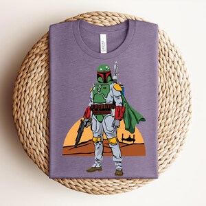 May include: A purple t-shirt featuring a graphic of a character in green and gray armor, a green cape, and a blaster. The design includes a sunset and a spaceship silhouette. The shirt is folded on a woven mat.