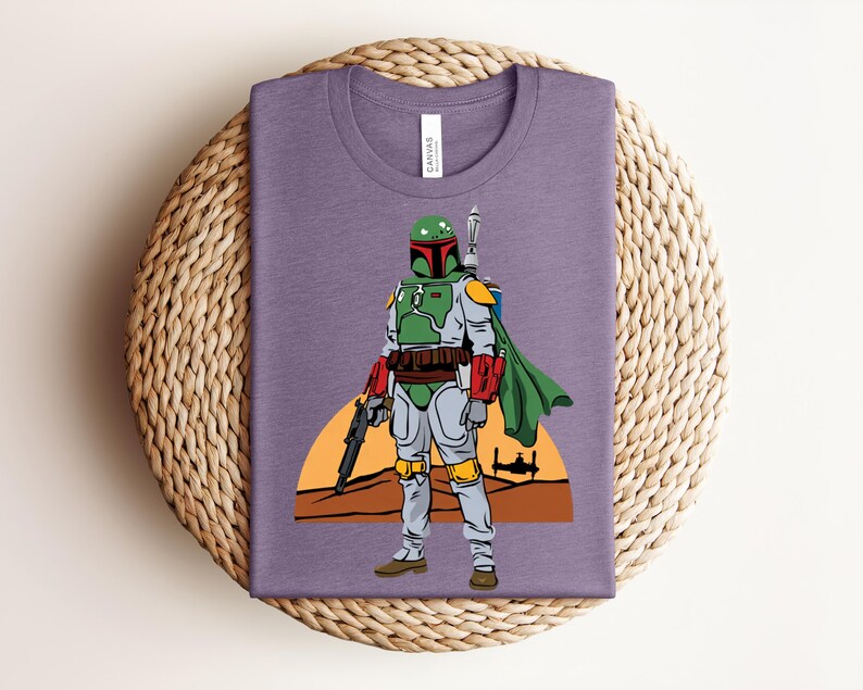 May include: A purple t-shirt featuring a graphic of a character in green and gray armor, a green cape, and a blaster. The design includes a sunset and a spaceship silhouette. The shirt is folded on a woven mat.