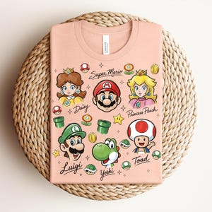 May include: A peach-colored t-shirt featuring Super Mario characters Daisy, Mario, Princess Peach, Luigi, Yoshi, and Toad. The shirt includes the words "Super Mario" and character names. The design incorporates stars, coins, and other game elements.