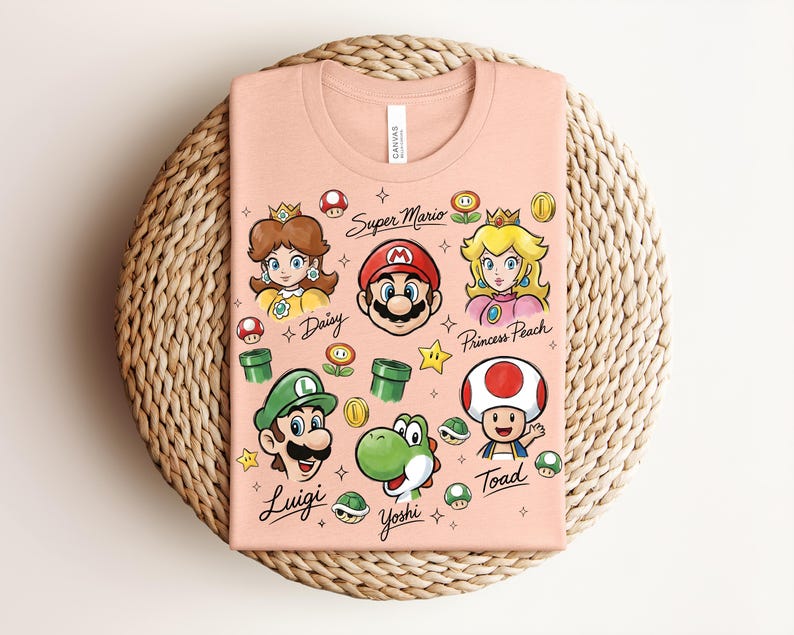 May include: A peach-colored t-shirt featuring Super Mario characters Daisy, Mario, Princess Peach, Luigi, Yoshi, and Toad. The shirt includes the words "Super Mario" and character names. The design incorporates stars, coins, and other game elements.