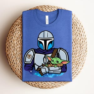 Puede incluir: Una camiseta azul con una ilustraci&oacute;n de dibujos animados de un personaje con armadura plateada que sostiene una peque&ntilde;a criatura verde. El dise&ntilde;o incluye un casco, una armadura para los hombros y una capa. La peque&ntilde;a criatura tiene orejas grandes y est&aacute; envuelta en una manta marr&oacute;n.