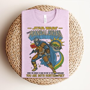 Puede incluir: Una camiseta de color morado claro con un estampado gr&aacute;fico de The Mandalorian y Grogu. El texto dice STAR WARS THE MANDALORIAN, BOUNTY HUNTER y WHEN YOU CHOOSE TO WALK THE WAY OF THE MANDALORE YOU ARE BOTH HUNTER & PREY en amarillo y azul.