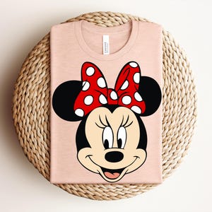 May include: A light pink t-shirt featuring a large graphic of Minnie Mouse's face. Minnie's face is black with a red and white polka dot bow. The shirt is folded and placed on a woven, circular surface.