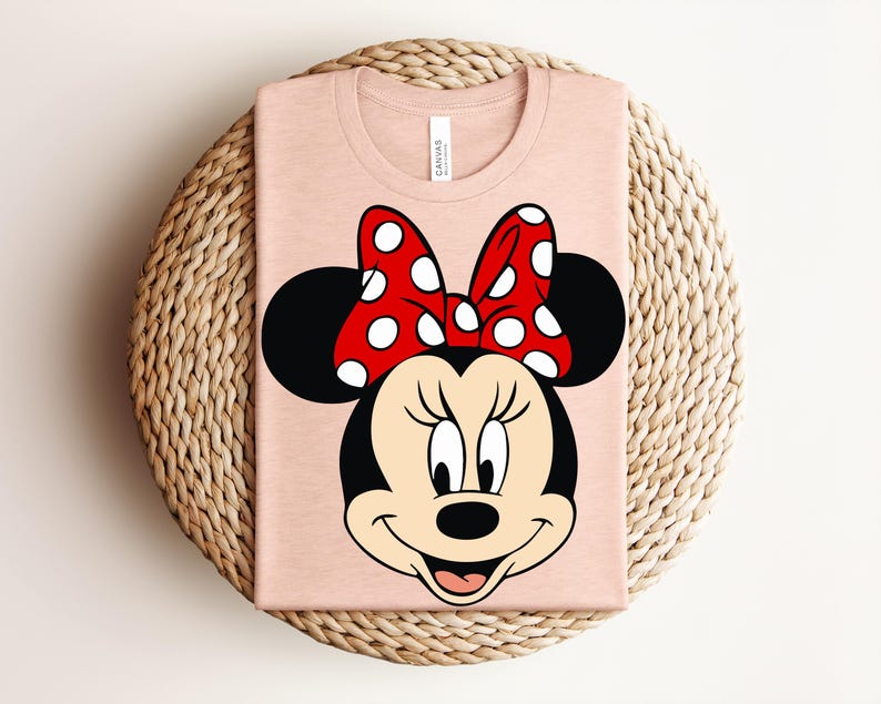 May include: A light pink t-shirt featuring a large graphic of Minnie Mouse's face. Minnie's face is black with a red and white polka dot bow. The shirt is folded and placed on a woven, circular surface.