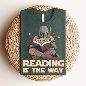 May include: A dark green t-shirt featuring a graphic of a helmeted character in armor sitting cross-legged and reading a book with a small green creature. The text below the graphic reads READING IS THE WAY in large, bold, light-colored capital letters.