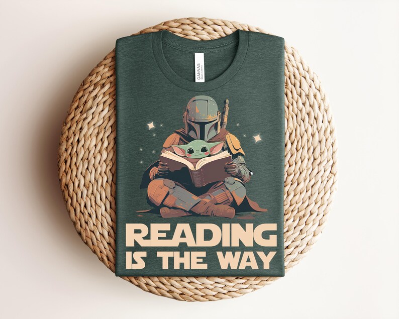 May include: A dark green t-shirt featuring a graphic of a helmeted character in armor sitting cross-legged and reading a book with a small green creature. The text below the graphic reads READING IS THE WAY in large, bold, light-colored capital letters.