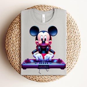 May include: A light gray t-shirt featuring a vibrant graphic of Mickey Mouse as a DJ. Mickey is wearing headphones and operating a turntable with blue and purple accents. The shirt is folded on a woven, circular surface.