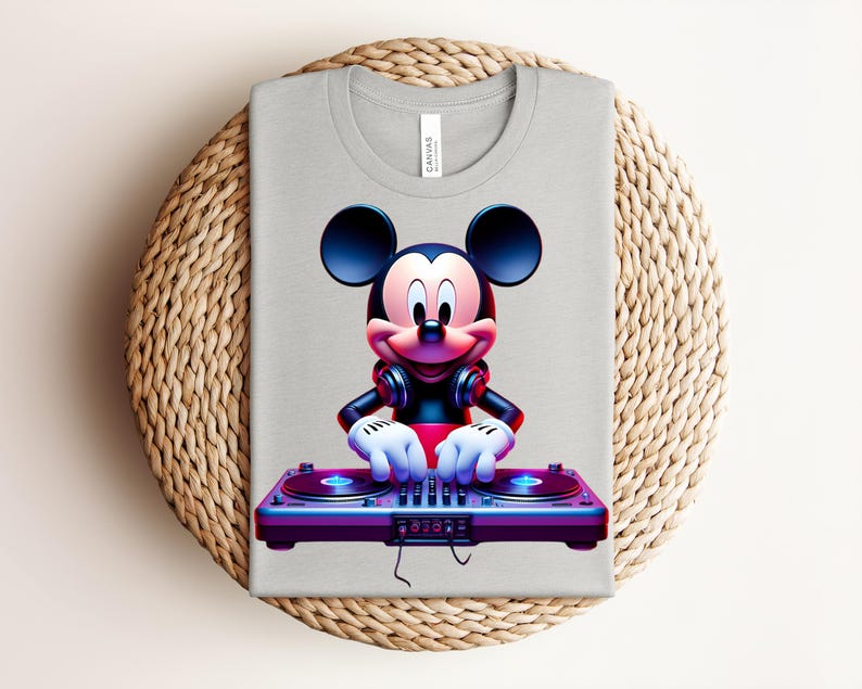 May include: A light gray t-shirt featuring a vibrant graphic of Mickey Mouse as a DJ. Mickey is wearing headphones and operating a turntable with blue and purple accents. The shirt is folded on a woven, circular surface.