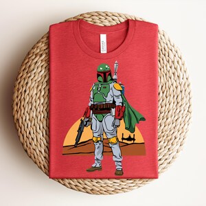 May include: Red t-shirt featuring a graphic of a character in green and gray armor, holding a weapon, with a green cape. The design includes a sunset and a spaceship silhouette.