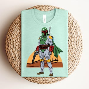 May include: A light green t-shirt featuring a cartoon illustration of a character in green and gray armor, holding a blaster. The design includes a desert landscape with a setting sun and a spaceship. The shirt is folded on a woven mat.