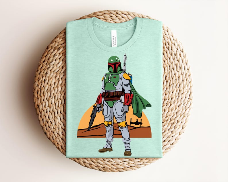 May include: A light green t-shirt featuring a cartoon illustration of a character in green and gray armor, holding a blaster. The design includes a desert landscape with a setting sun and a spaceship. The shirt is folded on a woven mat.