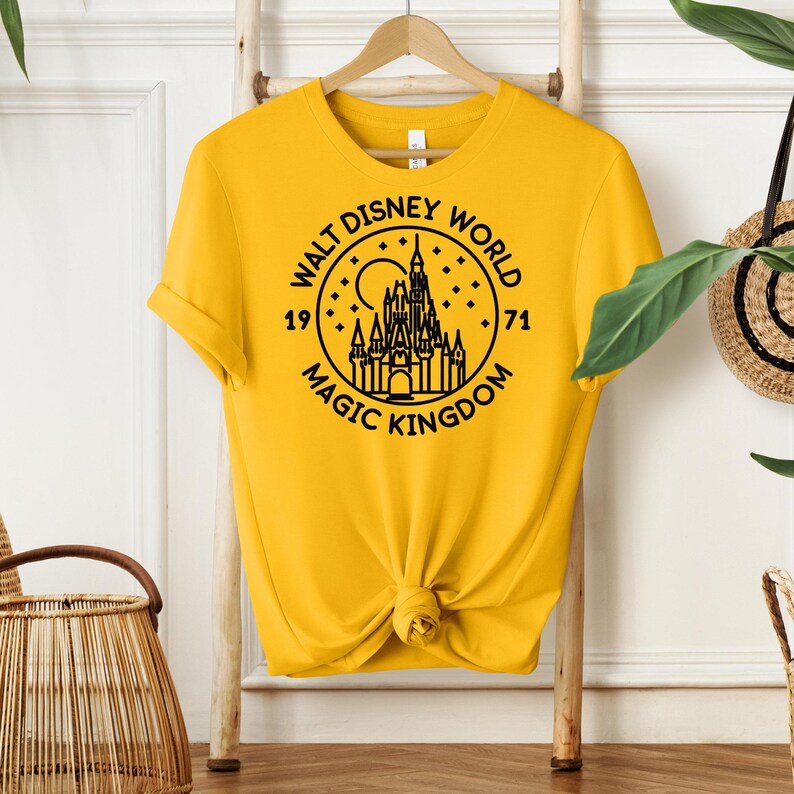 May include: A yellow t-shirt featuring a black graphic of the Walt Disney World logo with the words "Walt Disney World Magic Kingdom 1971". The shirt is knotted at the bottom.