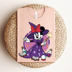 May include: A light pink t-shirt featuring a cartoon character dressed as a witch. The character wears a purple dress, a witch hat with a red bow, and holds a wand. The design includes potion bottles, a crystal ball, and open books.