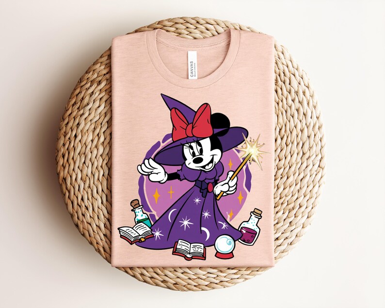 May include: A light pink t-shirt featuring a cartoon character dressed as a witch. The character wears a purple dress, a witch hat with a red bow, and holds a wand. The design includes potion bottles, a crystal ball, and open books.