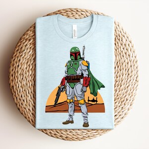May include: Light blue t-shirt featuring a cartoon illustration of a character in green and gray armor, holding a weapon. The character stands in front of a desert landscape with a setting sun and a spaceship. The shirt is folded on a woven surface.