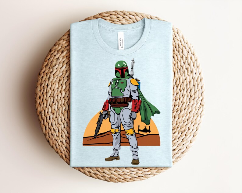 May include: Light blue t-shirt featuring a cartoon illustration of a character in green and gray armor, holding a weapon. The character stands in front of a desert landscape with a setting sun and a spaceship. The shirt is folded on a woven surface.
