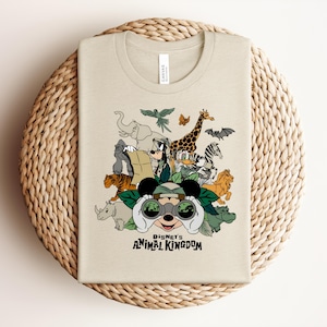 Puede incluir: Una camiseta de cuello redondo color beige con un estampado gr&aacute;fico de personajes de Disney, incluyendo a Mickey Mouse mirando a trav&eacute;s de binoculares, Goofy y el Pato Donald, rodeados de animales de safari como un elefante, una jirafa y un tigre con el texto Disney's Animal Kingdom.