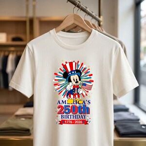 May include: Off-white t-shirt featuring a graphic of Mickey Mouse dressed in patriotic attire, holding an American flag. The design includes the text "America's 250th Birthday 1776-2026" in red, white, and blue colors. The shirt is hanging on a wooden hanger.