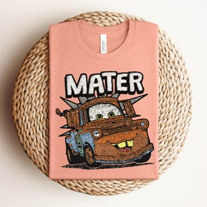 May include: A peach-colored t-shirt featuring a graphic illustration of the character Mater from the movie Cars. The design includes the name MATER in large white letters above the rusty brown tow truck, which has green eyes and a wide smile.