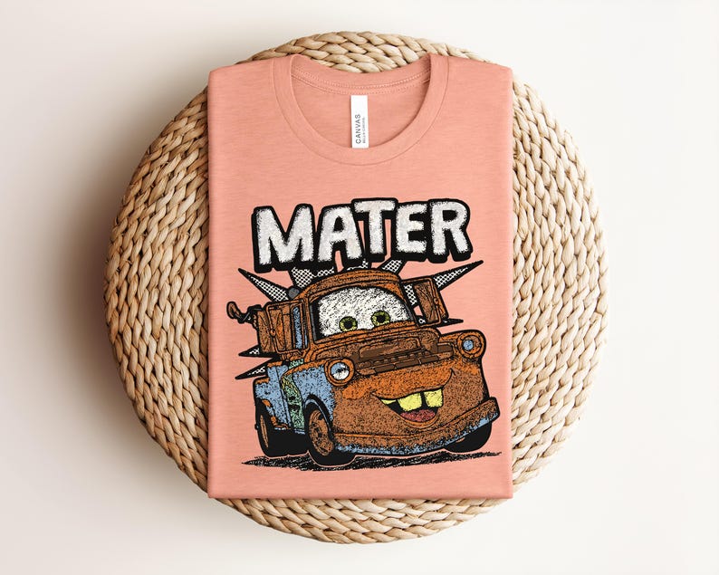 May include: A peach-colored t-shirt featuring a graphic illustration of the character Mater from the movie Cars. The design includes the name MATER in large white letters above the rusty brown tow truck, which has green eyes and a wide smile.