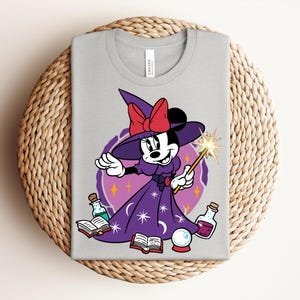 May include: A light gray t-shirt featuring a cartoon Minnie Mouse dressed as a witch. She wears a purple dress, hat, and holds a wand. The design includes potion bottles, a crystal ball, and open books. A red bow adorns her hat.