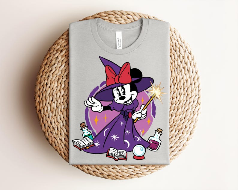 May include: A light gray t-shirt featuring a cartoon Minnie Mouse dressed as a witch. She wears a purple dress, hat, and holds a wand. The design includes potion bottles, a crystal ball, and open books. A red bow adorns her hat.