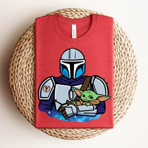 Puede incluir: Camiseta roja con una ilustraci&oacute;n de dibujos animados de un personaje mandaloriano sosteniendo a un personaje Grogu. El mandaloriano lleva un casco y una armadura, y Grogu es verde con orejas grandes. El dise&ntilde;o est&aacute; sobre un fondo espacial azul y morado.