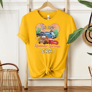 K&ouml;nnte beinhalten: Senfgelbes T-Shirt mit einer Grafik von Zeichentrickautos und dem Text "In my Radiator Springs ERA". Das Design umfasst Cartoon-Kakteen und Sterne. Das T-Shirt ist an der Taille geknotet und h&auml;ngt an einem Holzb&uuml;gel.