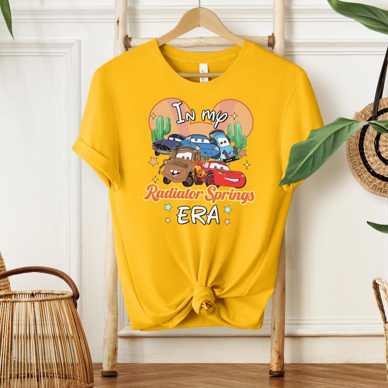 K&ouml;nnte beinhalten: Senfgelbes T-Shirt mit einer Grafik von Zeichentrickautos und dem Text "In my Radiator Springs ERA". Das Design umfasst Cartoon-Kakteen und Sterne. Das T-Shirt ist an der Taille geknotet und h&auml;ngt an einem Holzb&uuml;gel.